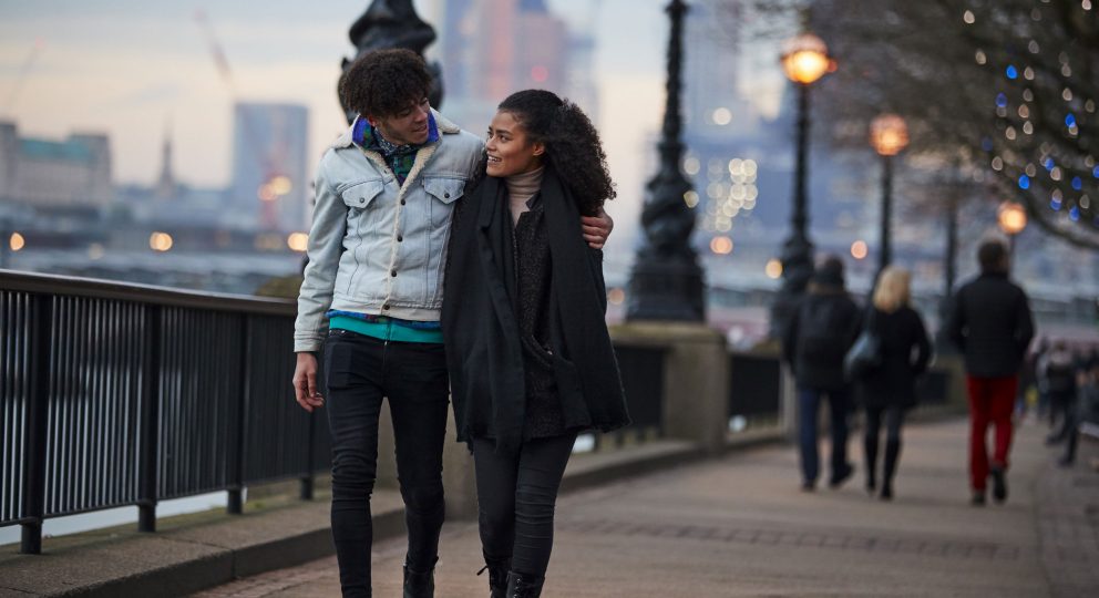 A couple walks down a boardwalk. The partner on the left has his arm slung over their partner.