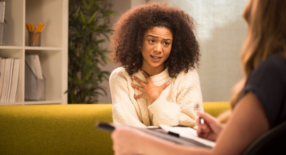 A photo of a woman sitting on a green couch from the point-of-view of a therapist's seat. The woman is talking and gesturing at herself.