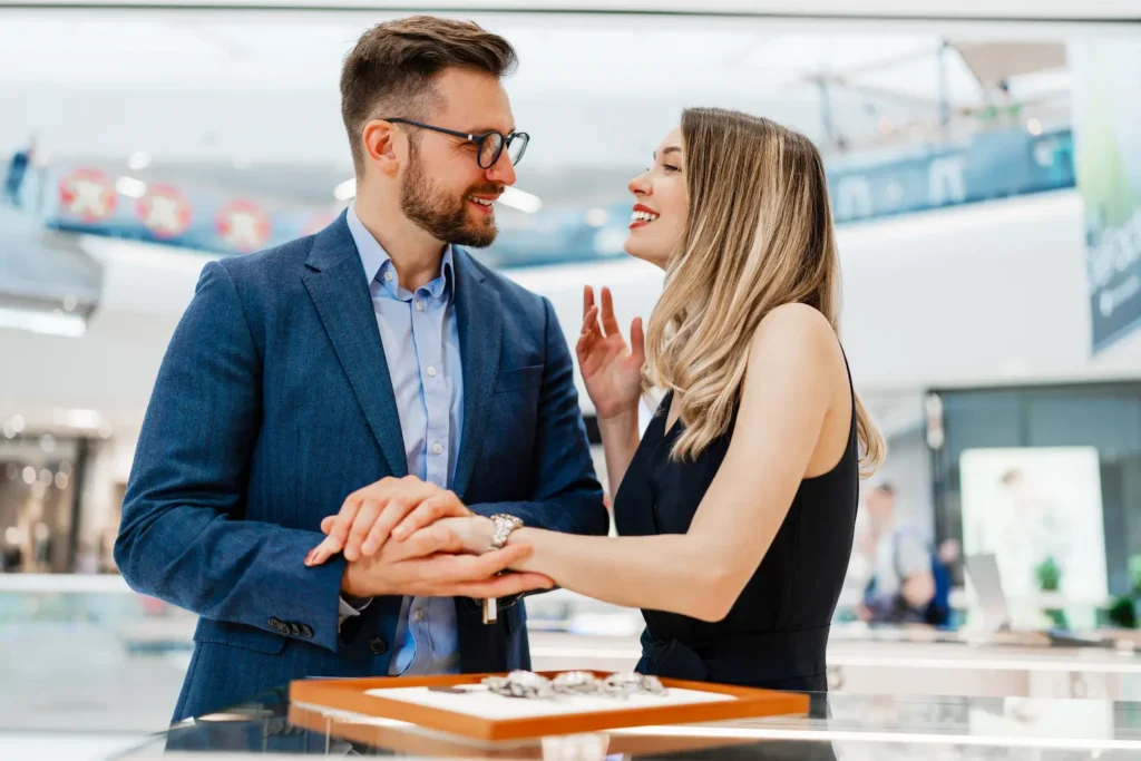 A well-dressed couple in an upscale jewelry store, with the man in a blue suit gazing intently at a smiling woman while holding her hand over an expensive jewelry display - potentially illustrating love bombing through lavish gift-giving and excessive attention early in a relationship.