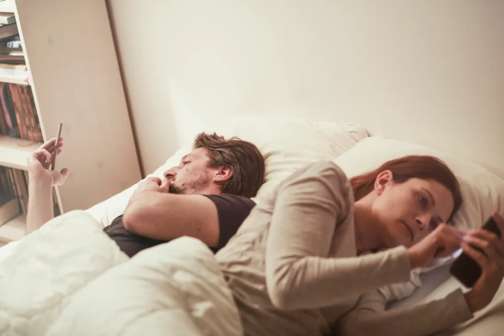 A couple lying in bed, both on screens- together physically but emotionally distant.